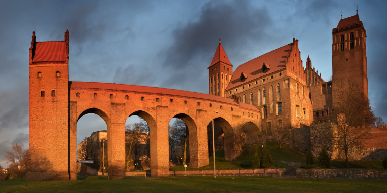 Beleuchtete Backsteinburg mit hohen Türmen, Kirche und arkadenartiger Brücke im warmen Abendlicht vor einem dunklen, wolkigen Himmel.