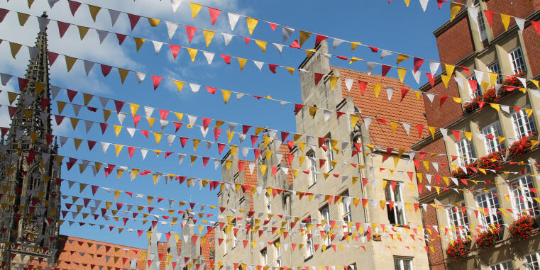 Numerous red, yellow, and white pennant strings (bunting) are stretched across the Prinzipalmarkt in Münster, Germany. In the background, the characteristic historic gabled houses and the spire of St. Lamberti Church are visible against a bright blue sky. Red geraniums bloom in the window boxes of one of the buildings.