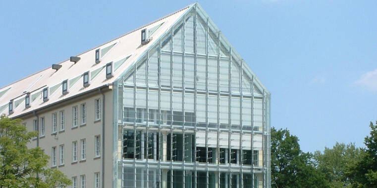 Ein modernes Gebäude mit einer großflächigen Glasfassade an der Giebelseite. Der angrenzende Gebäudeteil ist hell verputzt mit regelmäßigen Fensterreihen. Das Dach ist mit mehreren Dachfenstern versehen. Im Hintergrund sieht man grüne Bäume und einen klaren blauen Himmel.
