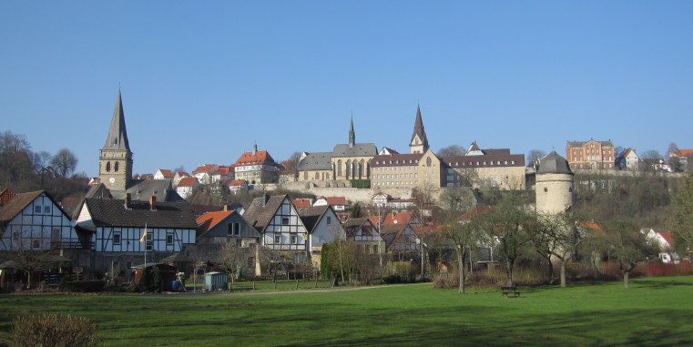 Blick über eine grüne Wiese auf eine Kleinstadt mit Fachwerkhäusern, mehreren Kirchen und einem runden Turm auf einer Anhöhe unter klarem blauem Himmel.