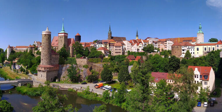 Panorama einer historischen Altstadt mit steinernen Wehrtürmen, Stadtmauer, roten Dächern und Kirchtürmen über einem Fluss, umgeben von viel Grün unter blauem Himmel.