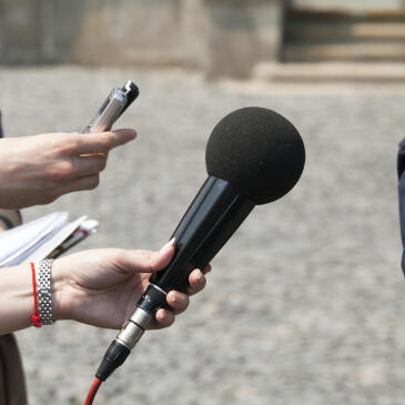 Hände halten ein Mikrofon und ein Aufnahmegerät in Richtung eines kaum sichtbaren Menschen am rechten Bildrand.