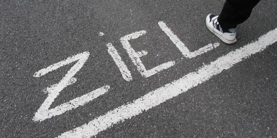 Mit weißen großen Buchstaben ist das Wort Ziel auf Asphalt geschrieben. Darunter ist eine Linie. Sie wird gerade von einem Menschen überquert, von dem nur Fuß und Unterschenkel zu sehen sind.