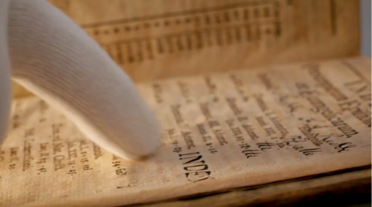 Zeigefinger in weißem Handschuh fährt über die Seite eines alten Buches.