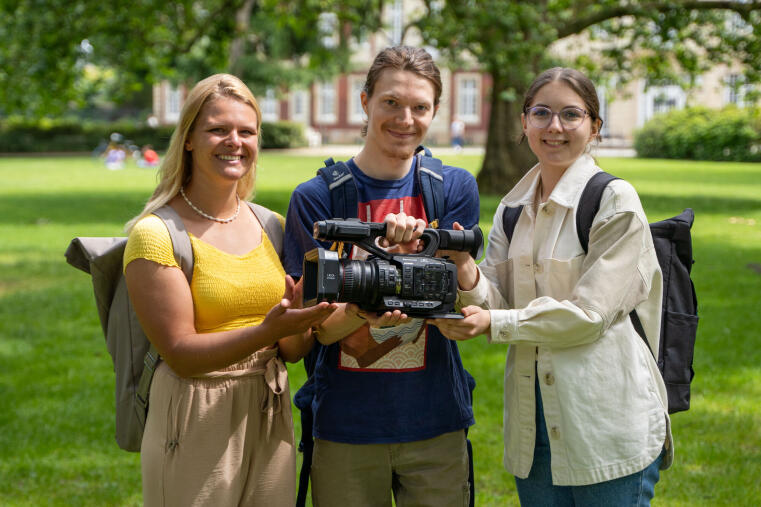 Das Bild zeigt 3 Personen im Schlossgarten, die gemeinsam eine Kamera halten.