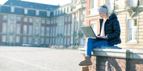 A woman sitting with a laptop. Behind is the facade of the University of Münster.