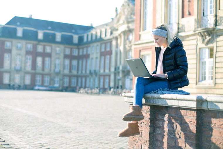 Im Vordergrund sitzt eine Frau mit einem Laptop auf dem Schoss. Im Hintergrund das Schloss der Universität Münster.