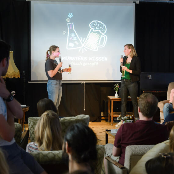 Foto einer Veranstaltungssituation: Zwei Menschen stehen vor Publikum auf der Bühne und interagieren miteinander.