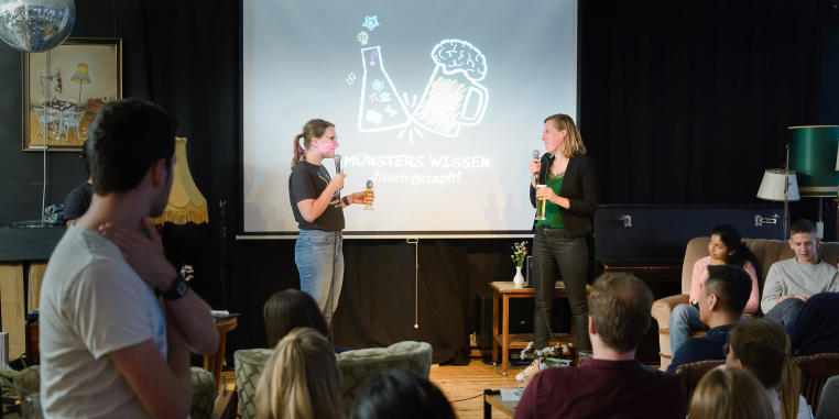 Picture of an event situation: Two people are standing on a stage in front of an audience and interacting with each other.