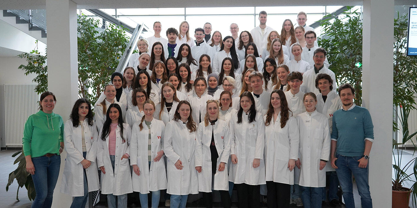Ein Gruppenbild: Alle Absolvent:innen des 25. PharMSchool-Durchgangs stehen im weißen Laborkittel auf der Treppe im Foyer des PharmaCampus, rehts und links eingerahmt vom Leitungsteam der PharMSchool.
