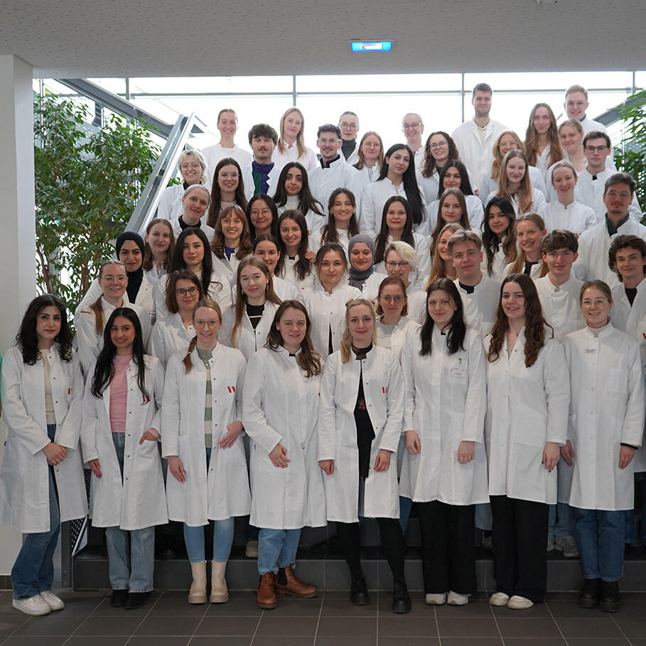 Ein Gruppenbild: Alle Absolvent:innen des 25. PharMSchool-Durchgangs stehen im weißen Laborkittel auf der Treppe im Foyer des PharmaCampus.