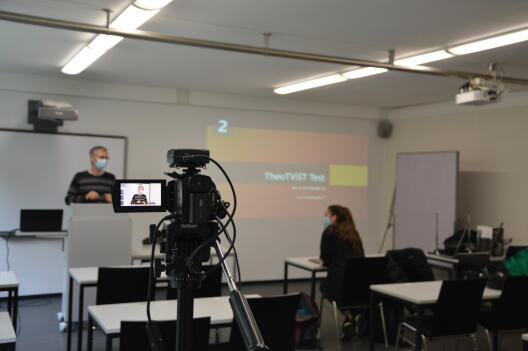 Foto von einer Kamera, die einen Seminarraum und eine vortragende Person filmt 