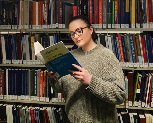 Auf dem Bild zu sehen ist eine Person mit etwas längeren, dunklen Haaren und einer Brille. Sie steht vor einem vollen Bücher-Regal in einer Bibliothek. In der Hand hält sie ein Buch mit dem Titel "Gedächtnis und Erinnerung. Ein interdisziplinäres Handbuch".