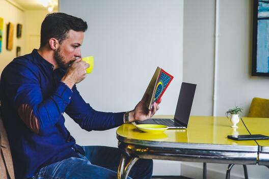 Männliche Person sitzt mit gelber Kaffetasse am Tisch und liest eine Zeitschrift, Auf dem Tisch steht ein augeklappter Laptop.zudem liegt ein Smartphone in Griffweite auf dem Tisch.
