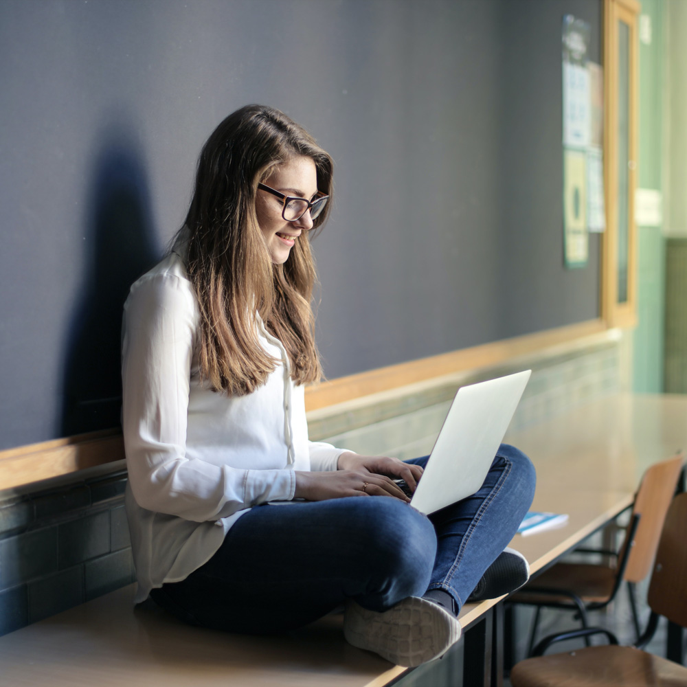 Eine junge Frau mit Brille sitzt im Schneidersitz vor einer Schultafel und hat auf dem Schoß einen Laptop, auf den sie interessiert blickt.