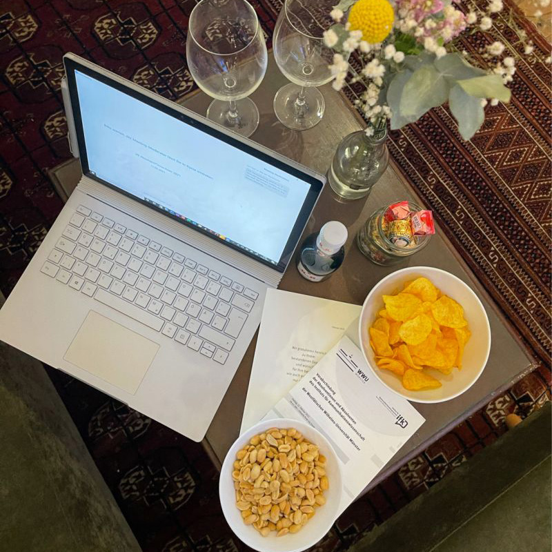 Ein Tisch, auf dem von oben fotografiert ein Laptop, zwei Schüsseln mit Chips und Erdnüssen, Sektgläser, eine Sektflsche, eine Vase mit Blumen sowie das Programmheft für die Feier liegen.