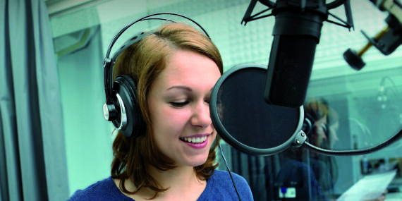 A female student talks into a studio microphone. She wears headphones. 