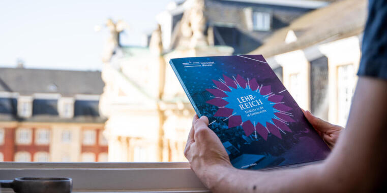Eine Person hält die gedruckte Publikation in den Händen, im Hintergrund ist das Schloss Münster zu sehen