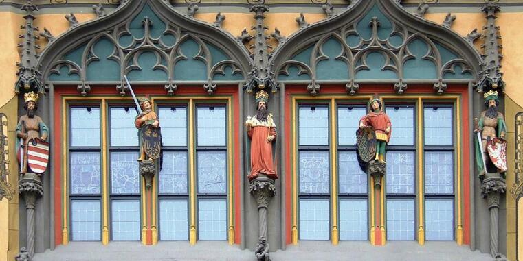 Zwei Sprossenfenster, außen stark verziert, an den Rahmen insgesamt fünf Statuen, einige davon mit Krone. Das Haus und die Verzierungen sind bunt. 
