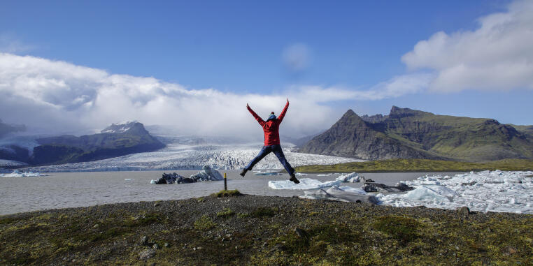 Eine Studentin macht einen Freudensprung vor der Gletscherlagune Fjallsárlón mit dem Fjallsárlón Gletscher im Hintergrund.