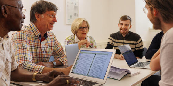 Ältere und jüngere Studierende sitzen im Seminar an einem Tisch und nutzen für ihre Gruppenarbeit mehrere Laptops.