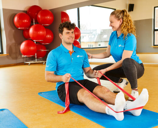 Junger Mann macht auf einer Matte eine Übung mit einem Fitnessband, daneben kniet eine Trainerin und unterstützt ihn.