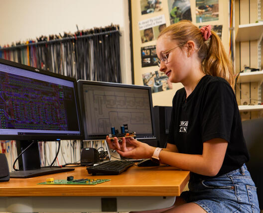 Eine junge Frau sitzt lächelnd an einem Schreibtisch mit zwei Bildschirmen und betrachtet eine elektronische Platine in ihrer Hand.