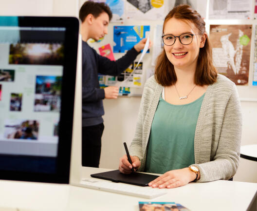 A young woman wearing glasses smiles while working with a graphics tablet at a computer, while a young man pins posters to a noticeboard in the background.