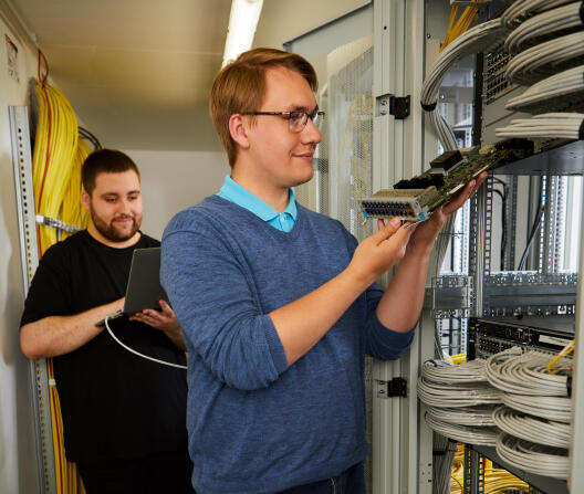 Zwei IT-Techniker arbeiten in einem Serverraum; einer prüft eine Netzwerkkarte, der andere bedient ein Laptop.