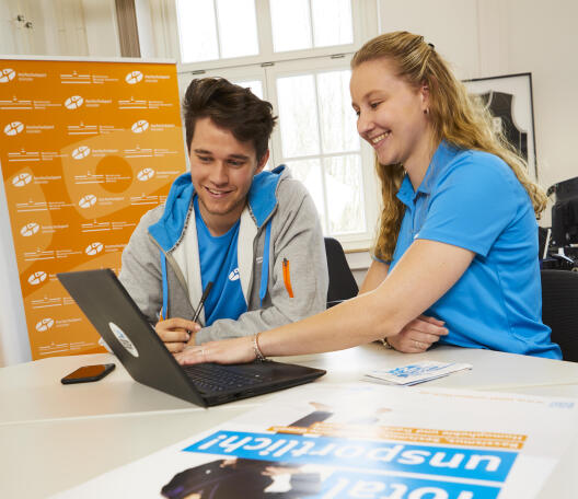 Ein junger Mann und eine junge Frau in blauer Hochschulsport-Kleidung sitzen lächelnd vor einem Laptop, im Hintergrund ein orangefarbenes Banner des Hochschulsports Münster.