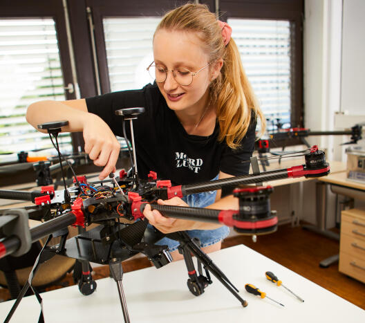 Eine junge Frau mit Brille arbeitet konzentriert an einer großen Drohne, daneben liegen Schraubendreher und ein robuster Laptop.