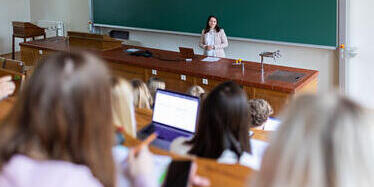 Students sit with laptops in a lecture hall, listening to a lecturer standing at the front desk.