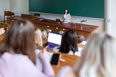 Lecturer giving a talk to students in a lecture hall with a green chalkboard; laptops and a tablet are visible on the desks in the foreground.