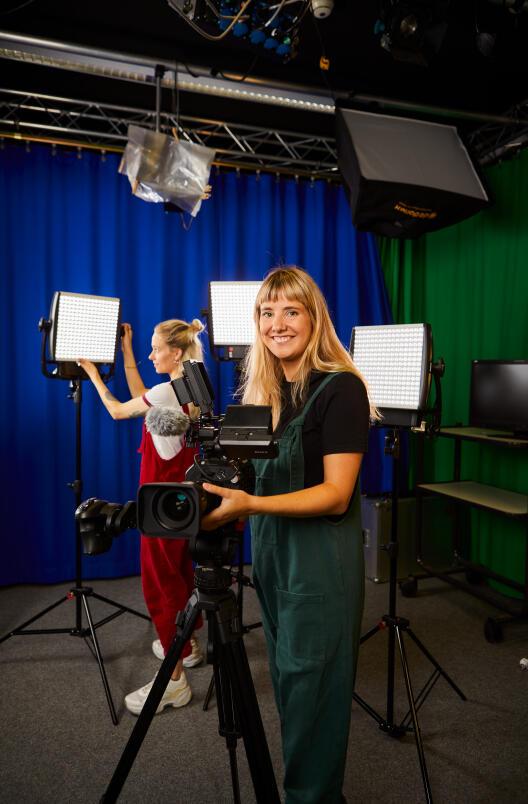A smiling young woman standing behind a professional video camera in a film studio, while a second person adjusts the lighting in the background.