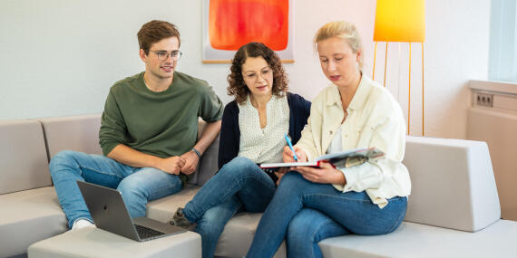 Three people sit together on a sofa, looking into a folder and discussing documents, with an open laptop beside them.