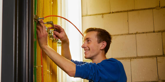 A young man in a blue jumper smiling while installing cables on a wall during a trade apprenticeship.