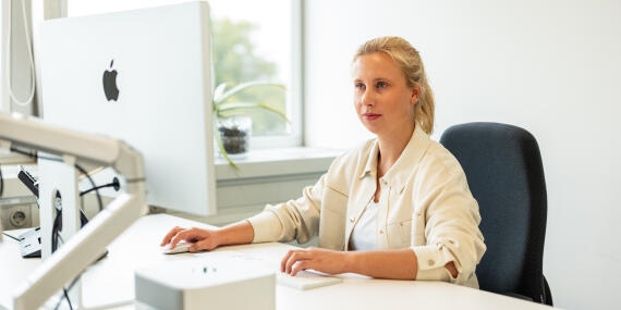 A young woman working concentratedly at a desk with an Apple computer in a bright office.