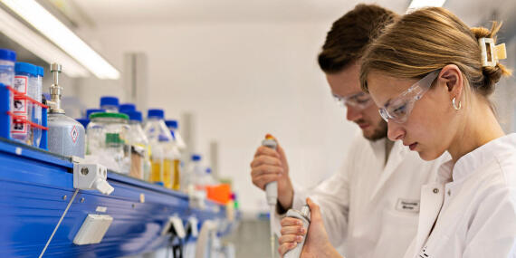 Two people in white lab coats and safety glasses work with pipettes in a laboratory next to a shelf of chemical bottles.