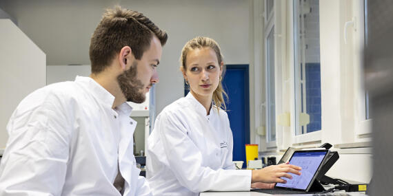 Two people in white lab coats work together on a tablet in a laboratory.