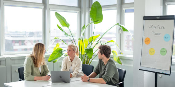 Three young persons having a discussion around a table with a laptop, next to a flip chart reading "Universität Münster".