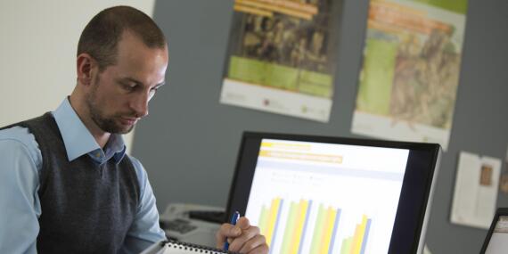 A man sits at a desk taking notes, with a screen showing a bar chart in the background.