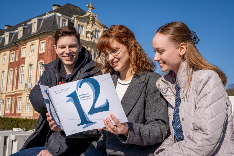 Drei Freunde schauen in die Publikation „Zwölf Monate, zwölf Menschen – Porträts 2025“. Sie sitzen auf einer Bank vor dem Schloss.