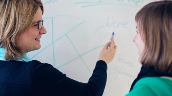 Two women are standing in front of a whiteboard. One woman draws something on the whiteboard and smiles as she explains something to the other woman.
