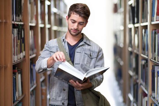 Junger Mann steht zwischen zwei Bücherregalen und blättert in einem Buch
