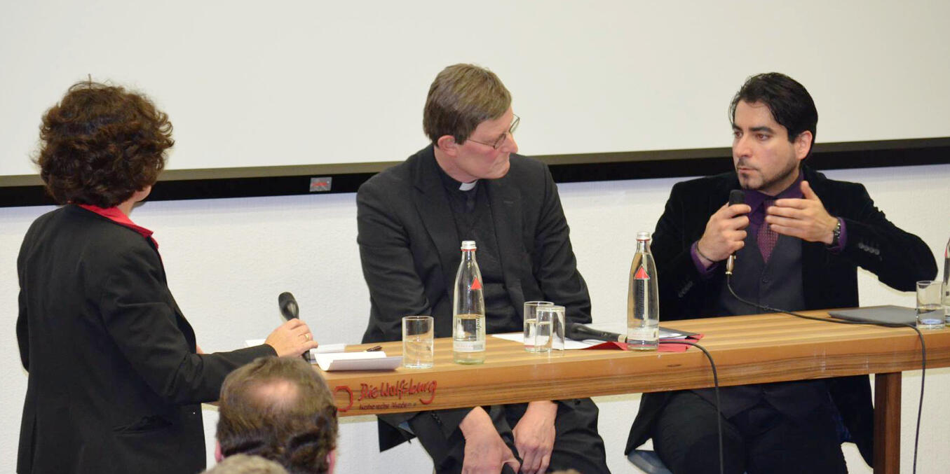 Rainer Maria Woelki und Prof. Dr. Mouhanad Khorchide sitzen hinter einem Tisch, eine Frau bringt gerade ein Mikrofon
