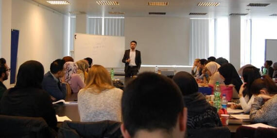 Dr. Çefli Ademi steht neben einem Whiteboard vor ihm mehrere Menschen an Tischen