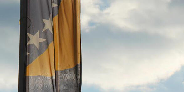 Himmel mit Wolken am linken Bildrand ist der untere Teil einer Fahne mit gelbem Aufdruck und gelben Sternen zu sehen