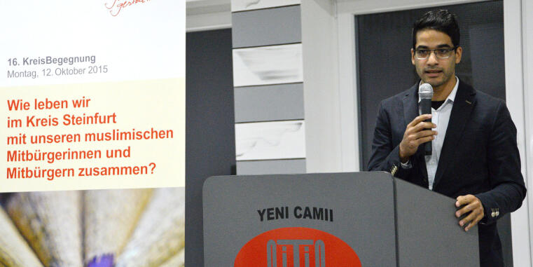 Dr. Amir Dziri hinter einem Rednerpult mit einem Mikrofon in der Hand, links befindet sich das Plakat der Veranstaltung