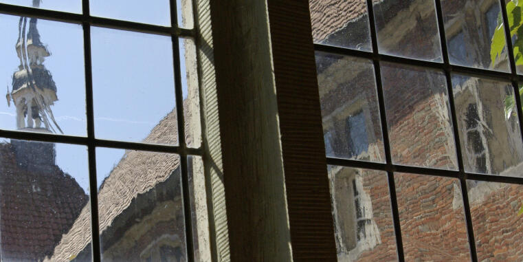 Sprossenfenster, durch welchees das Dach eines alten Gebäudes mit einem kleinen Turm zu sehen ist