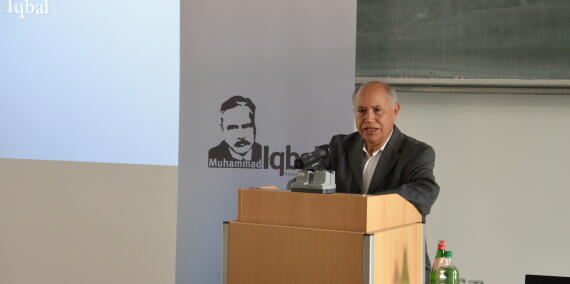Prof. Dr. Mohamed Turki hinter einem Rednerpult vor einem Plakat mit dem Konterfei Muhammad Iqbals